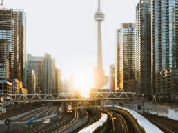 Three trains from the GO Transit network, Toronto's passenger rail system, pass by at sunrise. Skyscrapers are visible all around, with the CN Tower being the focal point.