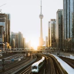 Three trains from the GO Transit network, Toronto's passenger rail system, pass by at sunrise. Skyscrapers are visible all around, with the CN Tower being the focal point.