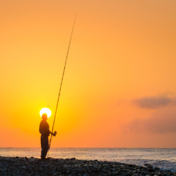 A local fisherman on the Taiwanese coast fishes at sunrise. The low sun illuminates the sky in a vivid orange colour.