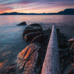 A vivid sunset is seen from one of Vancouver's rocky beaches. The tantalus mountain range is visible in the background.