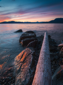A vivid sunset is seen from one of Vancouver's rocky beaches. The tantalus mountain range is visible in the background.