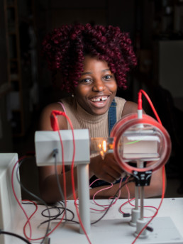 A young female engineering student smiles and laughs while she uses a piece of electrical engineering test equipment.