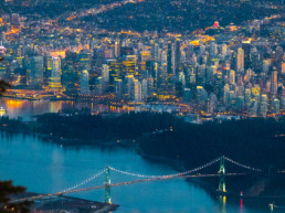 Vancouver's Lions Gate Bridge and the downtown core are visible at dusk from a viewpoint on the North Shore Mountains. The city's lights shine bright as the light in the sky fades. BC Place is visible in the background