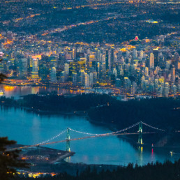 Vancouver's Lions Gate Bridge and the downtown core are visible at dusk from a viewpoint on the North Shore Mountains. The city's lights shine bright as the light in the sky fades. BC Place is visible in the background
