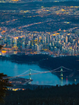 Vancouver's Lions Gate Bridge and the downtown core are visible at dusk from a viewpoint on the North Shore Mountains. The city's lights shine bright as the light in the sky fades. BC Place is visible in the background