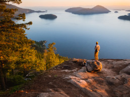 A hiker looks out at the view over Howe Sound from a viewpoint at the top of a hike. In the view is the ocean and a number of small islands. Golden sunlight makes the whole scene glow.