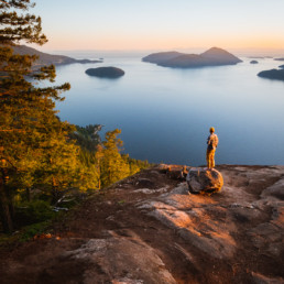 A hiker looks out at the view over Howe Sound from a viewpoint at the top of a hike. In the view is the ocean and a number of small islands. Golden sunlight makes the whole scene glow.