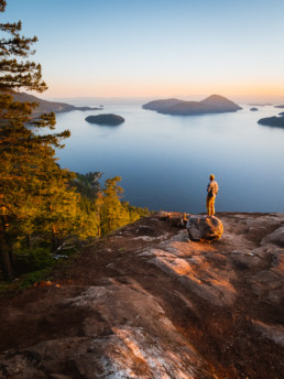 A hiker looks out at the view over Howe Sound from a viewpoint at the top of a hike. In the view is the ocean and a number of small islands. Golden sunlight makes the whole scene glow.