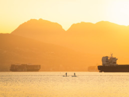 Two individuals paddleboard in Vancouver during the golden hour at sunrise. The sky is a bright orange colour. In the background, large ships are visible floating. Vancouver's north shore mountains are highlighted by the rising sun.