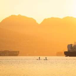 Two individuals paddleboard in Vancouver during the golden hour at sunrise. The sky is a bright orange colour. In the background, large ships are visible floating. Vancouver's north shore mountains are highlighted by the rising sun.