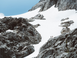Two hikers are seen from afar. They are silhouetted against a bright snowfield. A blue sky and the rocky peak are visible in the background.