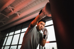 Young man punching a hanging boxing bag in a modern-style studio gym with large windows.